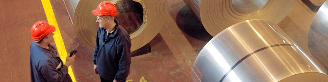 Two factory workers standing next to rolls of steel